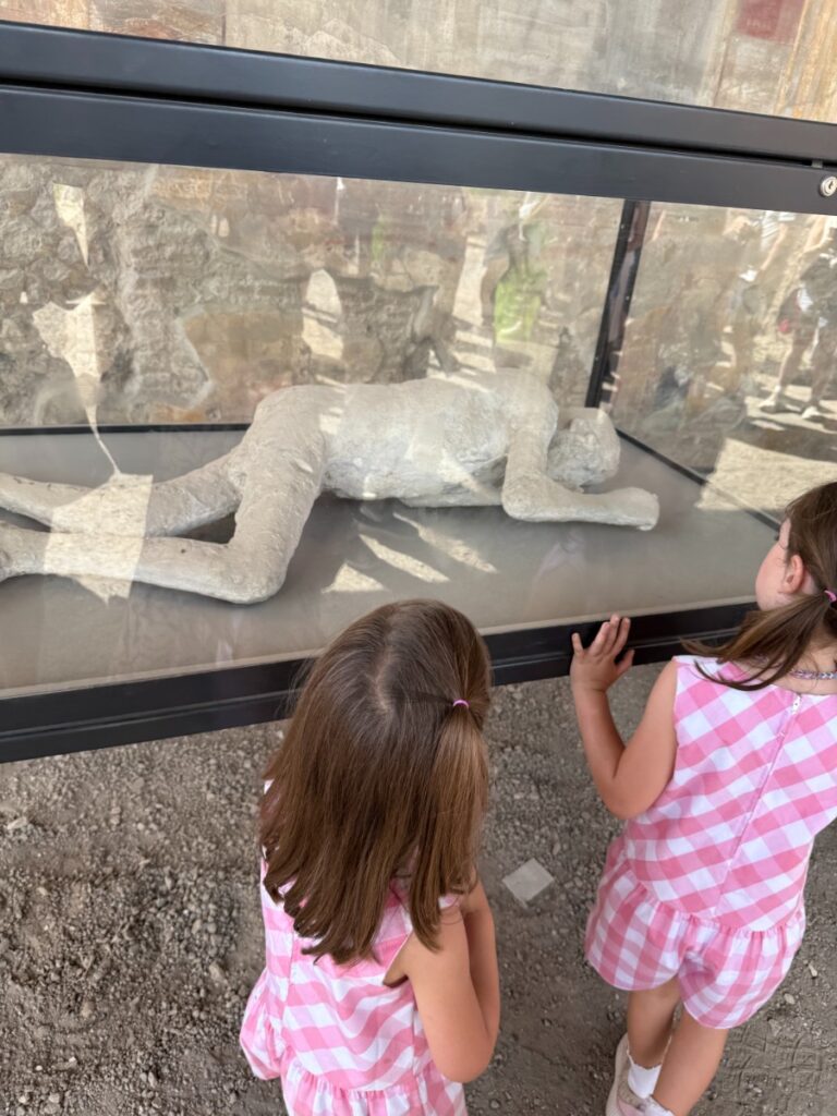 Two young girls in pink gingham dresses looking at a Pompeii body cast in a glass case during a family visit to the Amalfi Coast