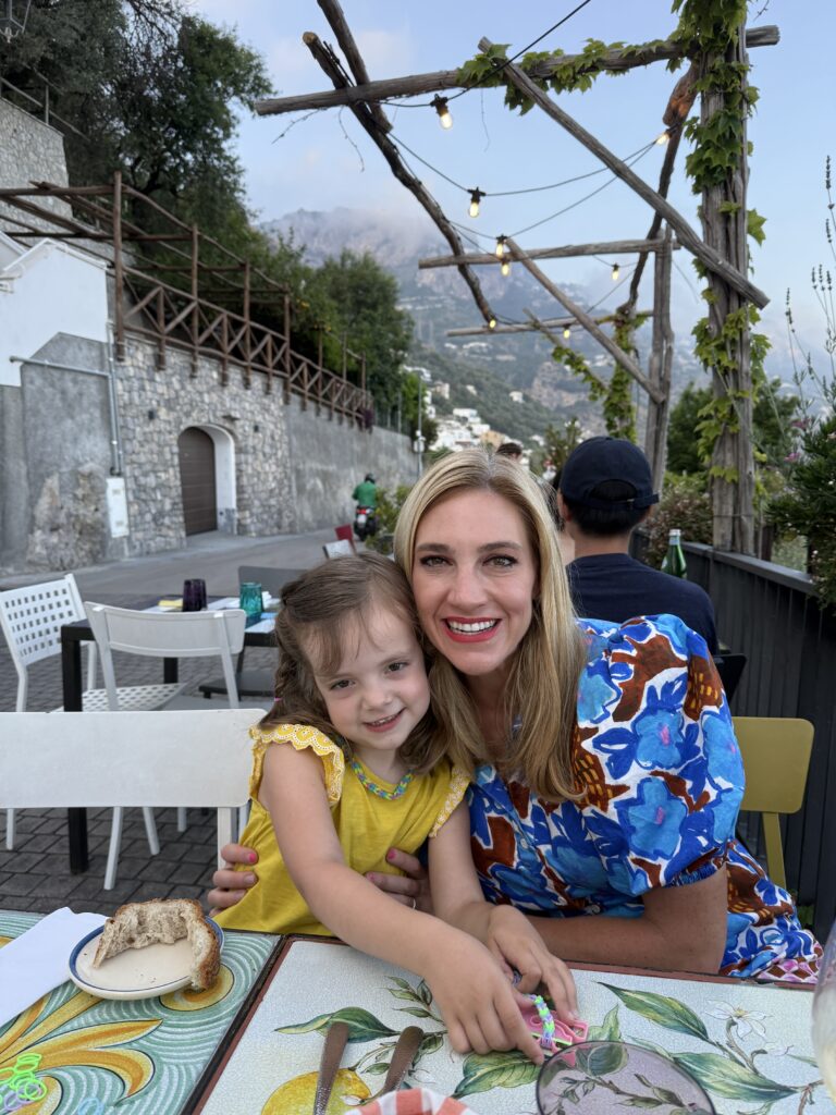 Mom and young daughter hugging at Kasai restaurant in Praiano on the Amalfi Coast with grape arbor, string lights, and mountains in the background